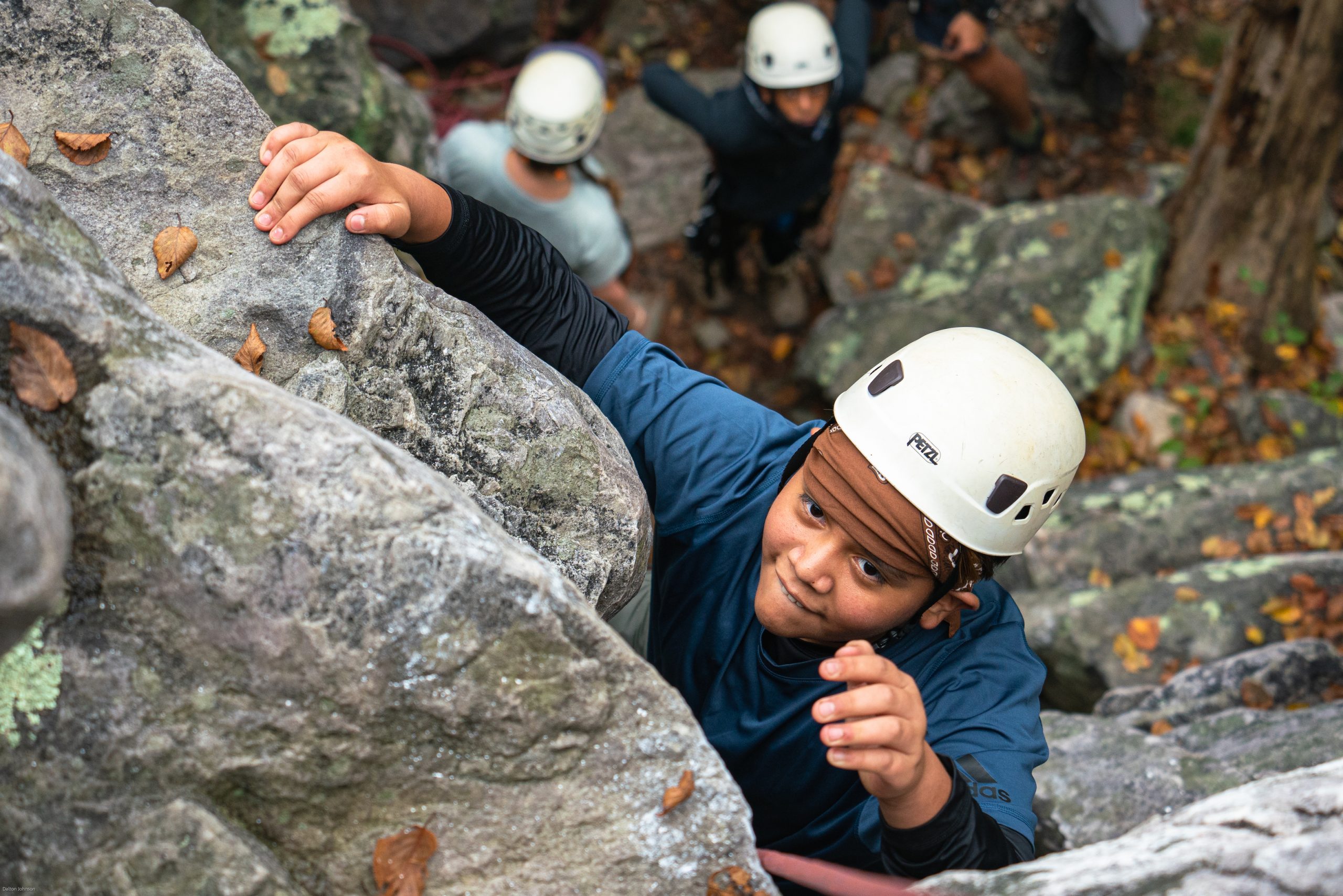 How Outward Bound is Different from Summer Camp: A student with a helmet on his reaching upward as they rock climb.