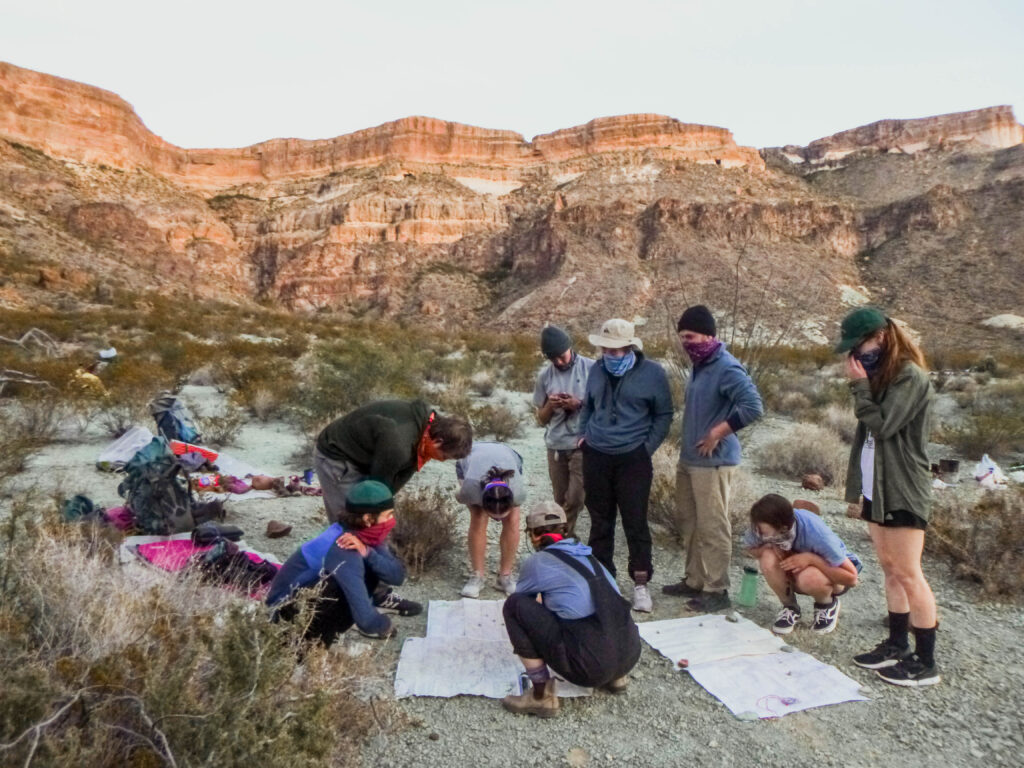 A group students in a desert gather around a map laying on the ground and utilize their strengths to make a plan. The sun is setting on the desert plateau behind them.