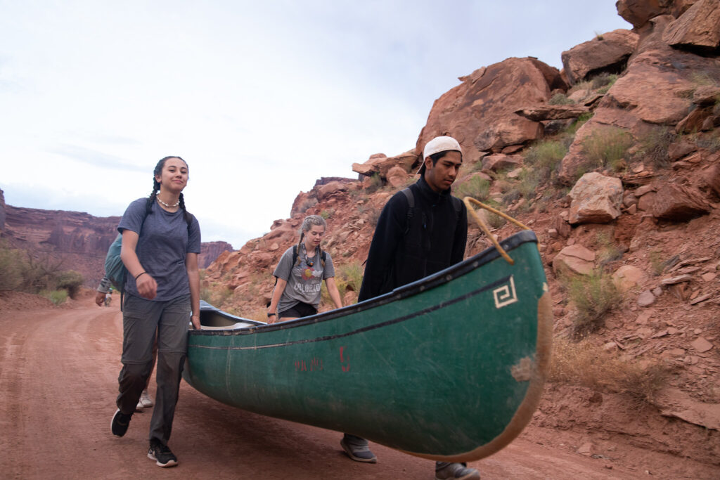 Three students carry a canoe up a red dirt path.