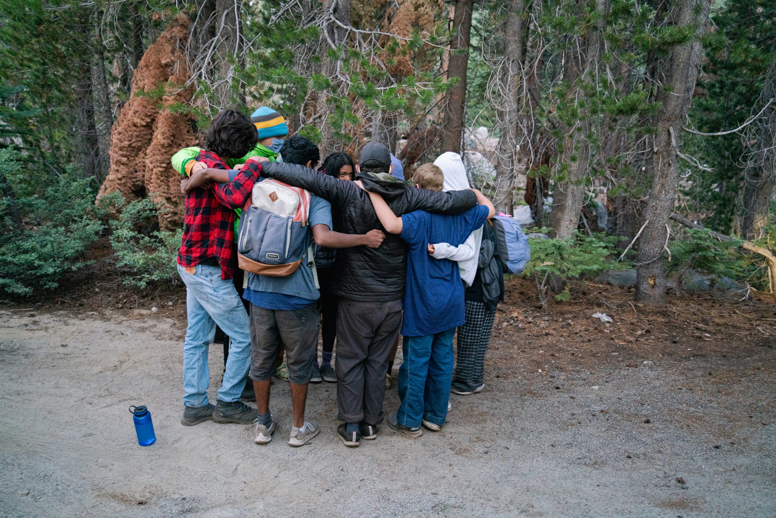 A group of individuals have a group hug outside with trees behind them.