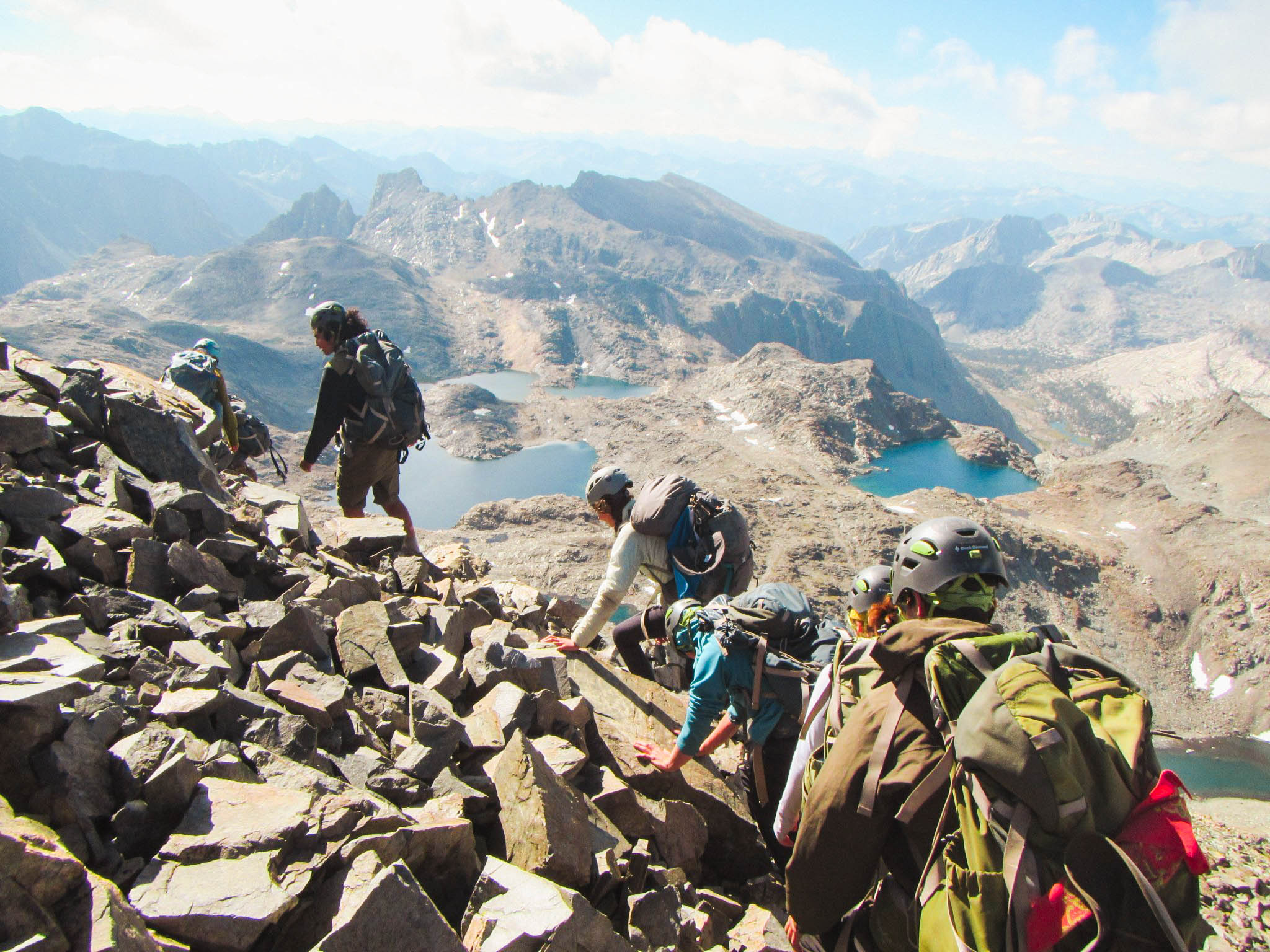 a group of six young adults with mountaineering helmets on and backpacks on scramble across rocks overlooking alpine lakes and mountains. 