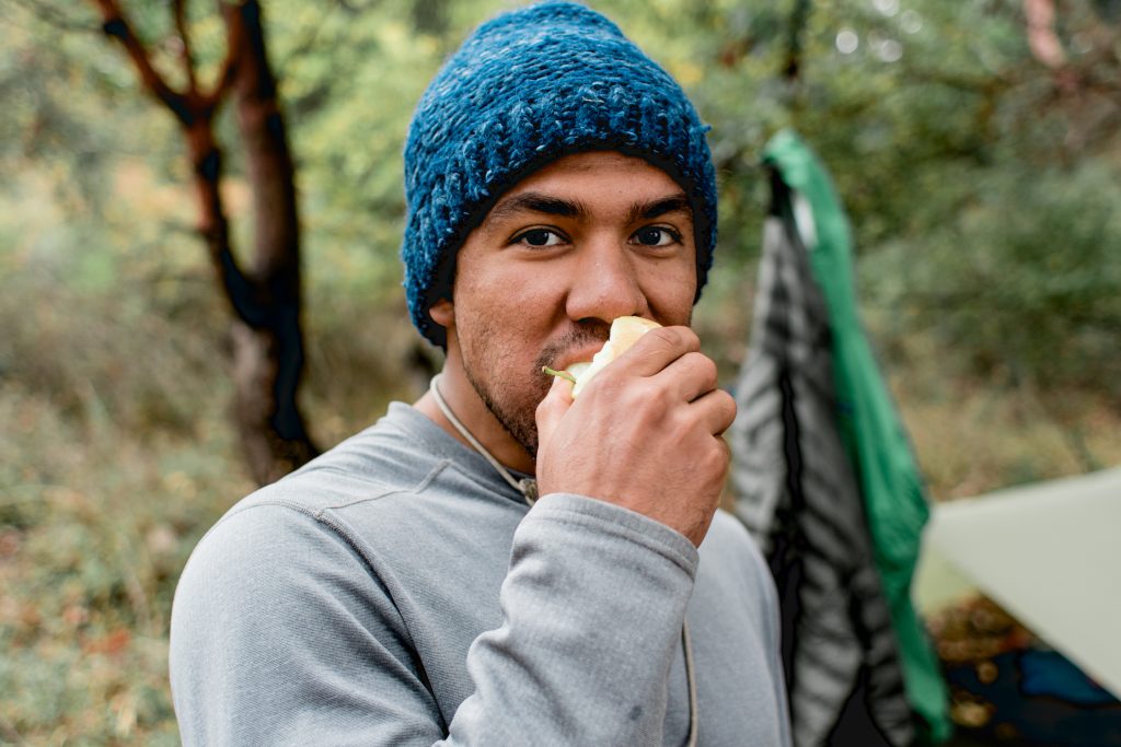  A person with a blue beanie on bites into an apple as they look into the camera. They are outside.