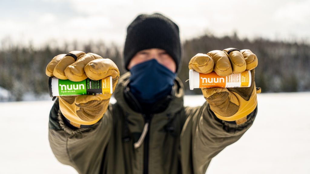 A person holds out in front of their face two Nuun Hydration packets in their hands. It is snowy in the background. hiking snacks blog photo.