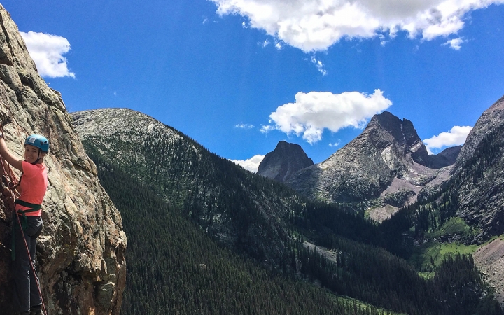 On the left side of the photo, a person wearing safety gear pauses during a rock climb. Behind them is a vast mountainous landscape under blue skies.