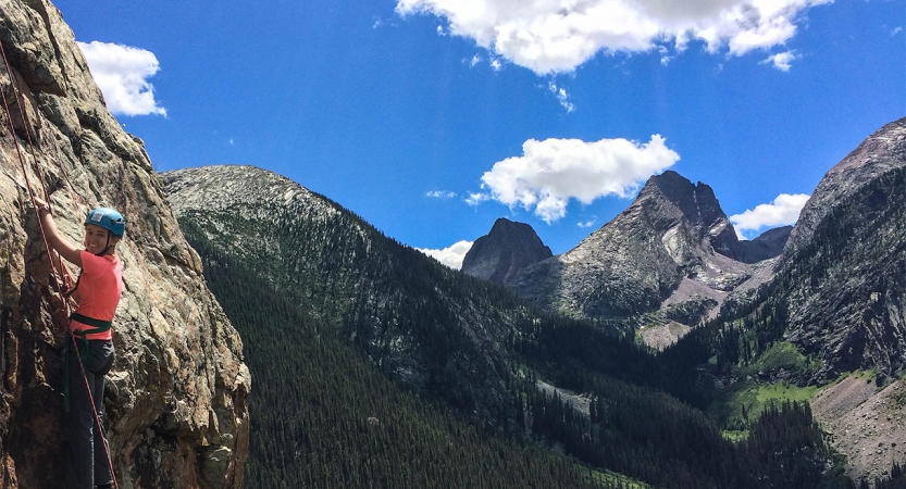 On the left side of the photo, a person wearing safety gear pauses during a rock climb. Behind them is a vast mountainous landscape under blue skies.
