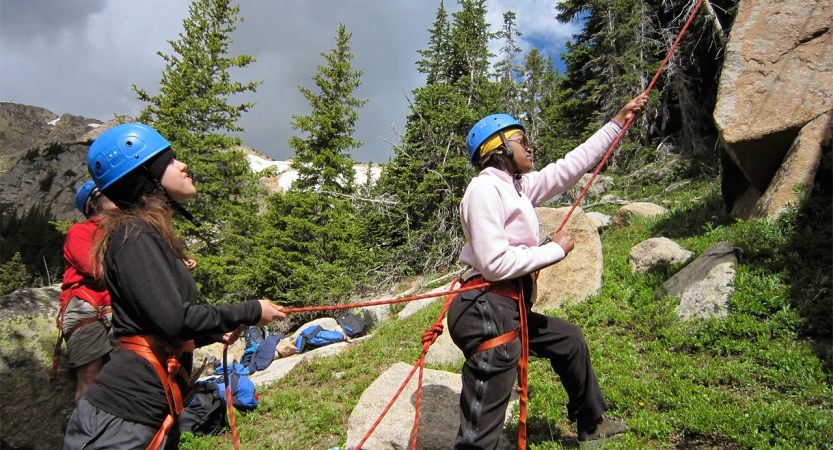 Three people wearing safety gear stand at the bottom of a rock wall and belay climbers.