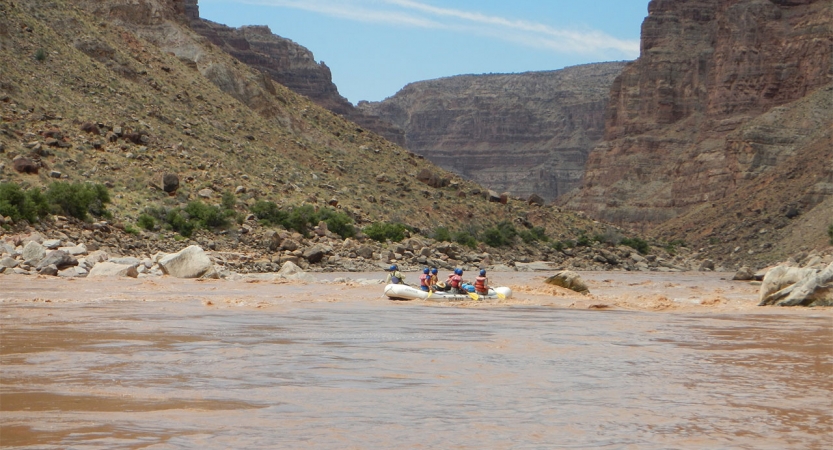 A group of students paddle a raft on a river framed by tall canyon walls