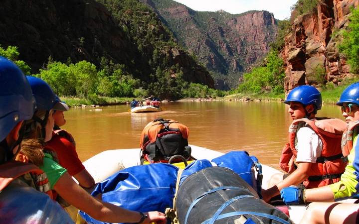 From the back of a raft, students are seen in front wearing safety gear. The water is calm, and there is another raft in the distance. The river is framed by tall canyon walls.