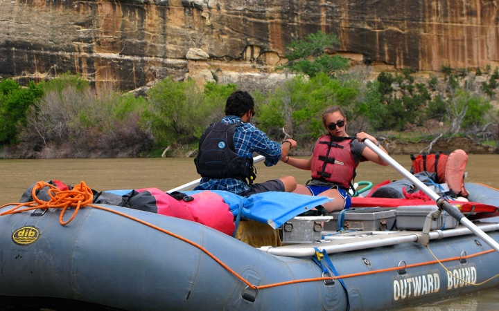 two people paddle a raft on calm water. behind them are tall canyon walls.