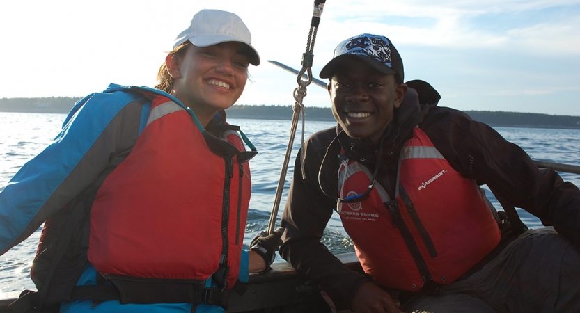 Two people wearing life jackets and hats smile while sitting inside a sailboat. 
