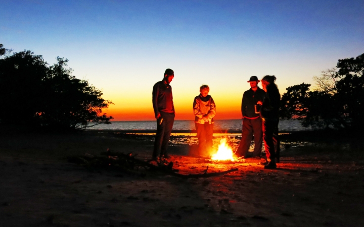 Four people stand around a campfire on a beach. The light appears in shades of blue, yellow and orange, as if the sun is setting or rising.
