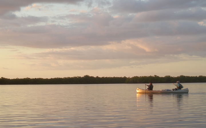 Two people paddle a canoe on calm water. The light appear soft, like the sun is setting or rising. There is a line of trees in the background.