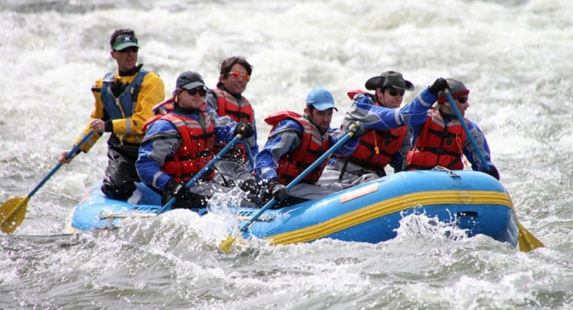A group of people wearing safety gear paddle a raft through whitewater.