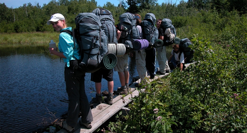 A group of students wearing backpacks stand on a narrow wooden plank over a body of water. Thick greenery surrounds the water. 