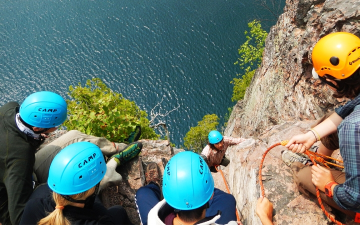 A group of students wearing helmets look over the edge of a cliff at a rock climber below. There is blue water below them.
