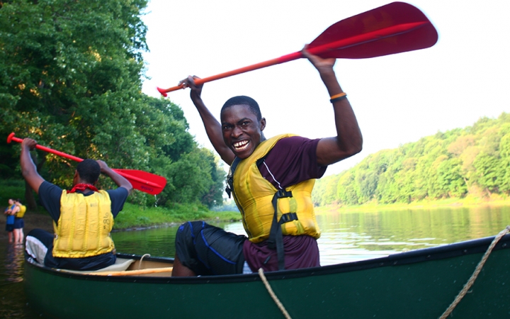 Two young people wearing life jackets sit in canoes and raise their paddles into the air in apparent celebration. One of them smiles at the camera. 