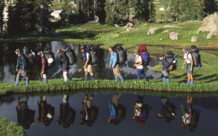 A group of people wearing backpacks hike on a grassy strip of land between two small bodies of water. Their images are reflected in the body of water in the foreground.