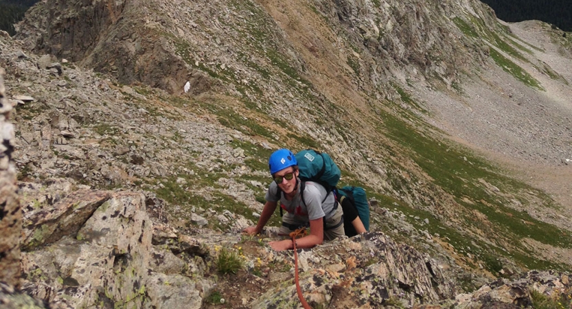 A person wearing safety gear looks up at the camera as they make their way up a rocky incline