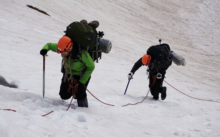 Two people wearing safety gear and carrying backpacks hold onto a rope as they ascend a snowy incline.