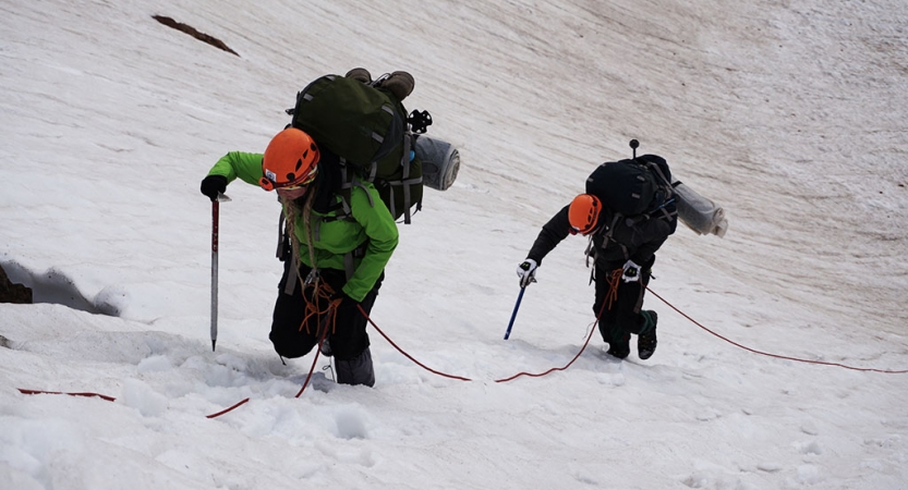 Two people wearing safety gear and carrying backpacks hold onto a rope as they ascend a snowy incline.