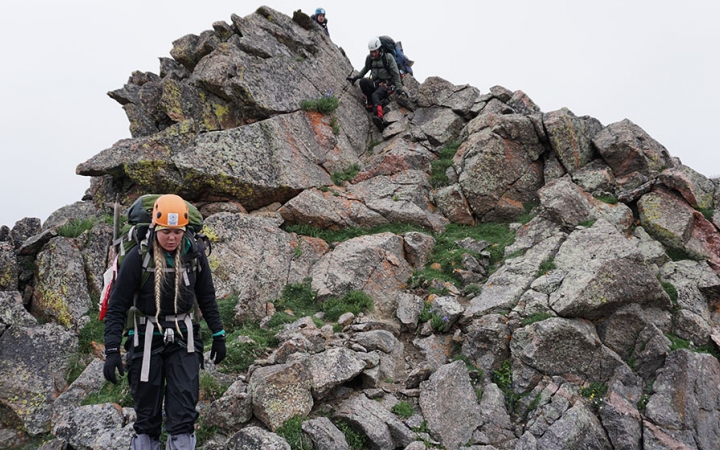 A person wearing a helmet carries a backpack as they hike near a rock formation.