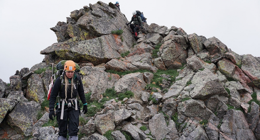 A person wearing a helmet carries a backpack as they hike near a rock formation.