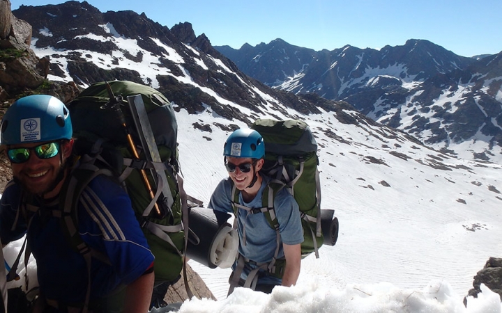 Two people wearing safety gear and backpacks smile as they hike along a snowy, mountainous landscape.