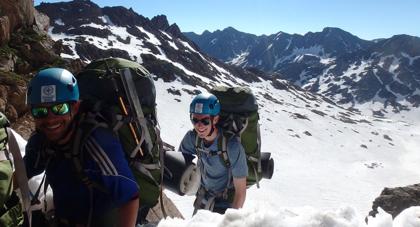 Two people wearing safety gear and backpacks smile as they hike along a snowy, mountainous landscape.
