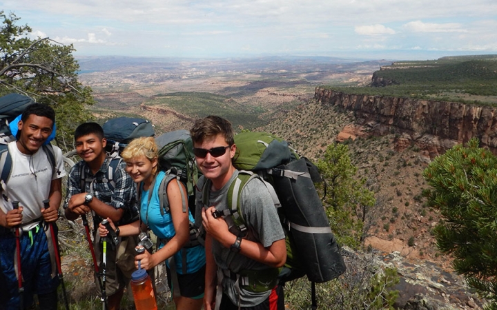 Four people wearing backpacks smile for a photo on an overlook high above a desert landscape.