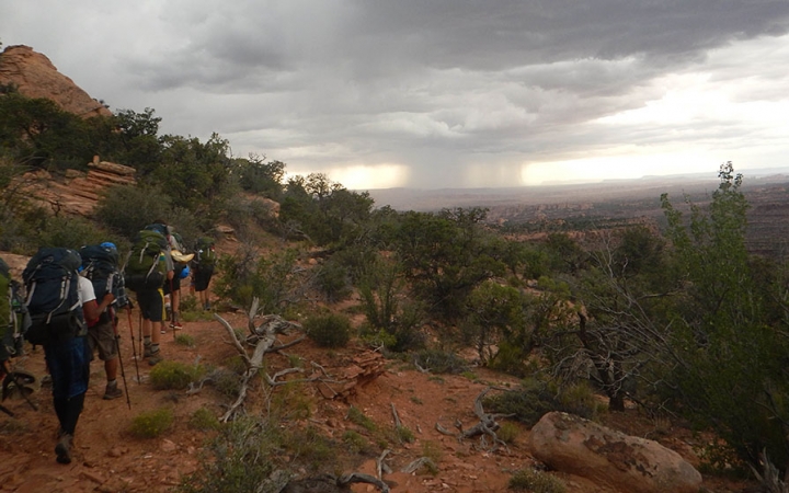 A group of people hike along a trail in a desert landscape. In the distance, it appears to be raining. 