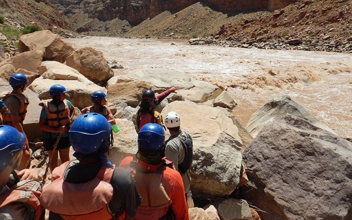 A group of people wearing helmets listen to an instructor as they provide instructions while pointing out to the river.