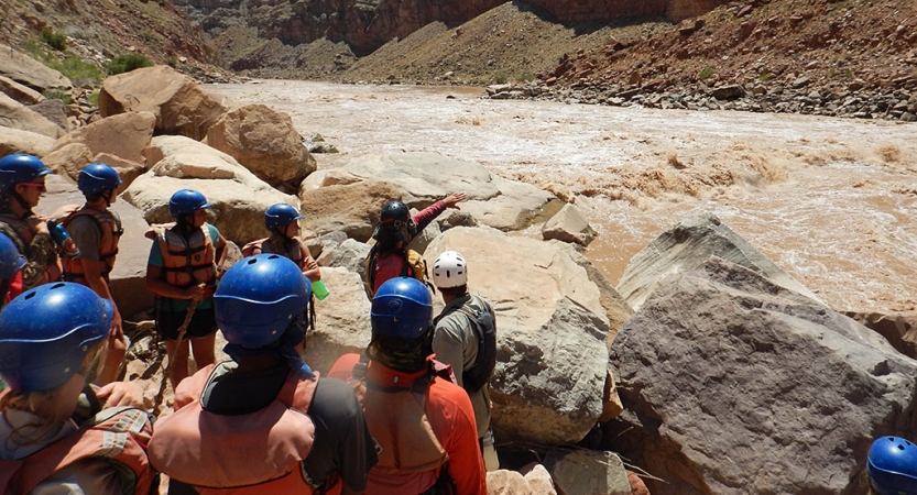 A group of people wearing helmets listen to an instructor as they provide instructions while pointing out to the river.