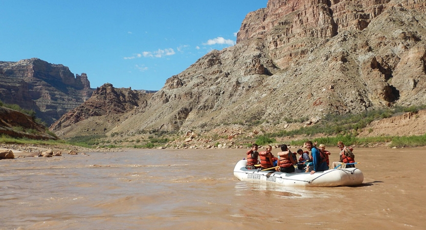 A group of people navigate a raft on a river sided by tall canyon walls