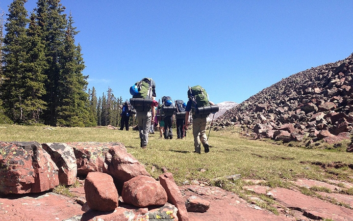 A group of students wearing backpacks high through a green meadow away from the camera 