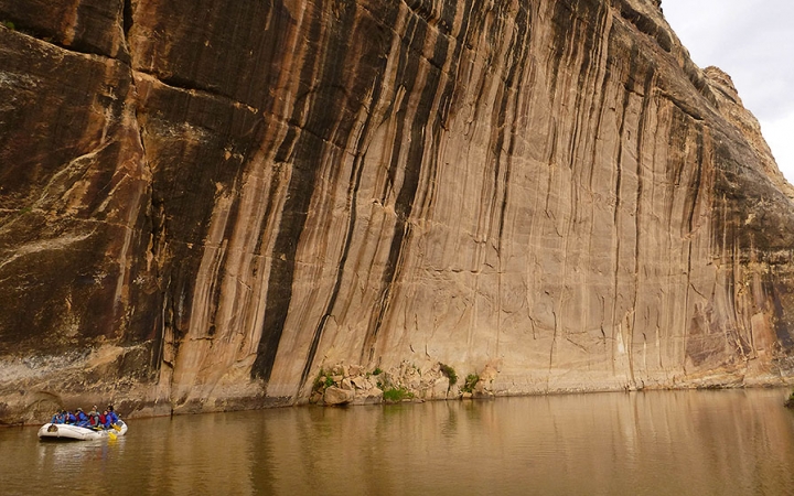 A raft is navigated on calm water beside a very steep and tall canyon wall.