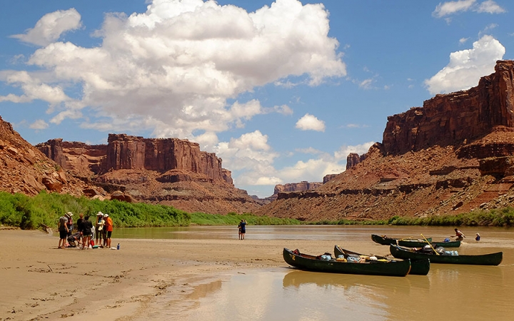 Three canoes rest on the shore of a river as a group of people stand nearby. There are tall, read mesas behind them.
