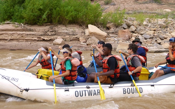 A group of students wearing safety gear paddle a green raft on a river. 