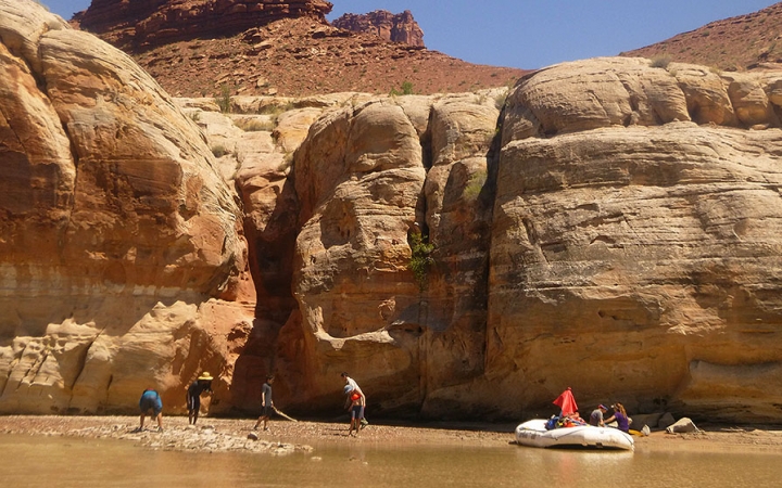 A raft is beached on a sandy shore, framed by tall canyon walls. There are people standing on the shore, too. 
