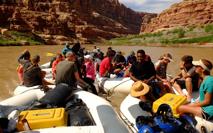 Four rafts are tethered together on the water. There are students sitting in each raft.
