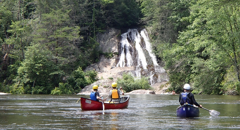 Two canoes are paddled by people wearing safety gear on calm water, toward a waterfall surrounded by trees.
