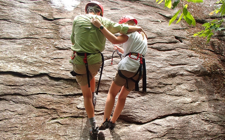 A parent and child wrap their arms around each other as they rappel down a rock wall. Both are wearing safety gear and are secured by ropes.