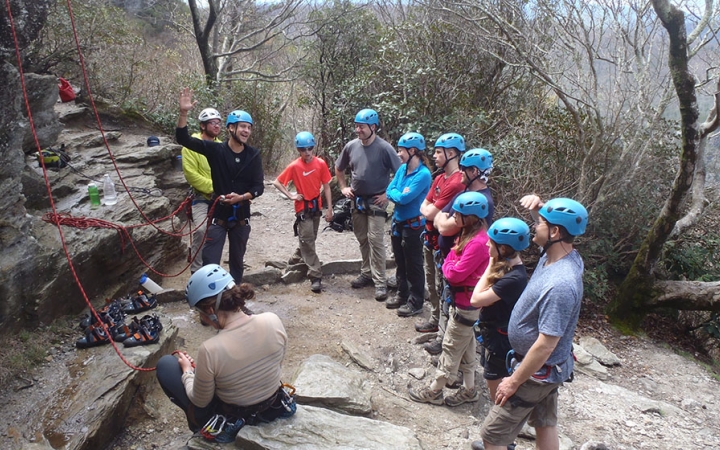 A group of people wearing safety gear stand in a line, listening to an instructor speak. They are standing near a rock wall, with ropes hanging down.