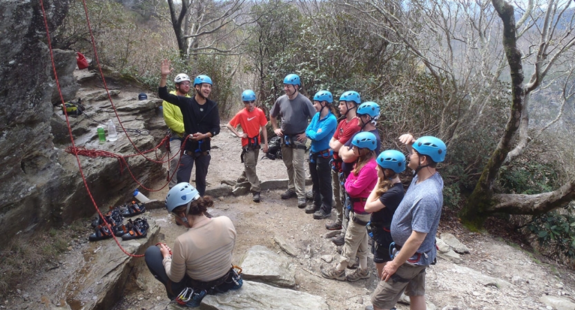 A group of people wearing safety gear stand in a line, listening to an instructor speak. They are standing near a rock wall, with ropes hanging down.