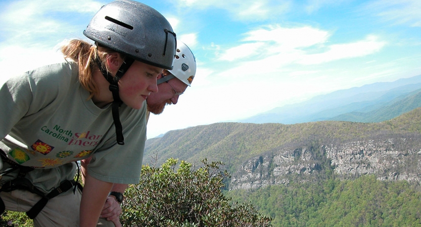 A parent and child wearing helmets look down over a vast mountainous landscape.