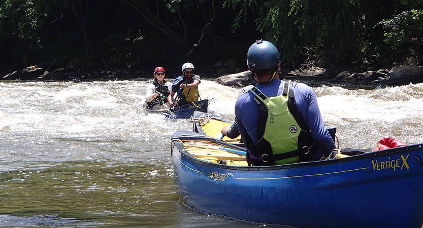 Two students paddle a canoe through whitewater, while another person who is likely an instructor watches them from another canoe.