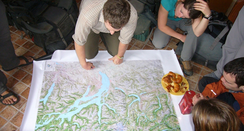 A map is spread out on the floor while a group of people examine it. 