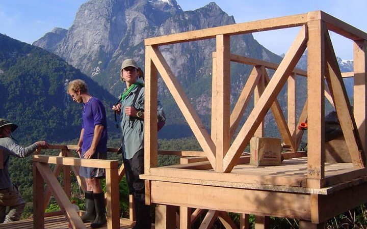 Three people appear to be working on the frame of a wooden structor. There is a tall mountain in the background. 