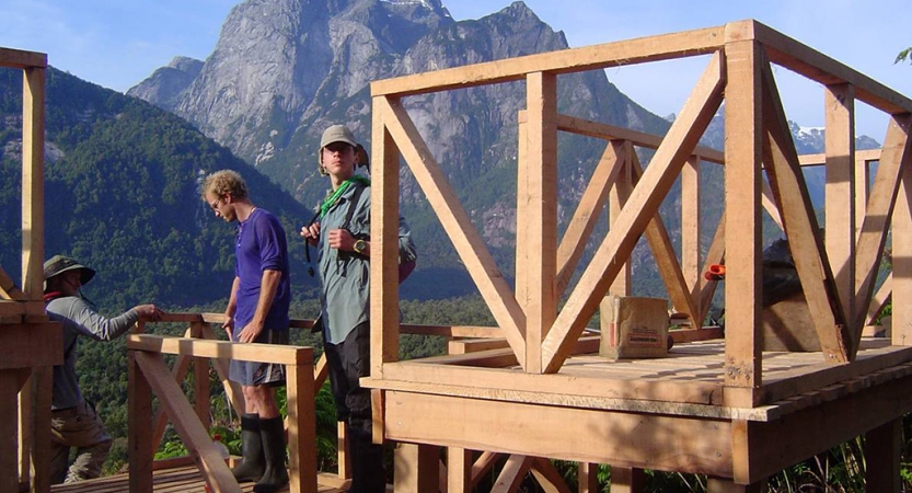 Three people appear to be working on the frame of a wooden structor. There is a tall mountain in the background. 