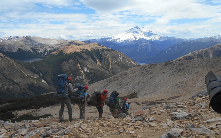 A group of people wearing backpacks hike along a rocky landscape. There is a vast mountainous landscape in the distance.