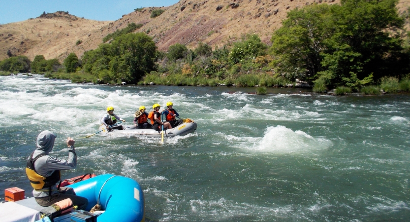 In the foreground, a person paddles a raft and watches another raft, behind them, being paddled by a group of students. 